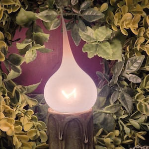 May include: A glowing, teardrop-shaped light bulb with a visible filament sits atop a dark green, textured candle holder. The candle holder has a dripped wax design. The background is filled with green and yellow foliage.