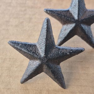 May include: Two black, cast iron star-shaped decorative objects. The stars have a textured surface and are set against a neutral background. These rustic stars could be used for home decor or craft projects.