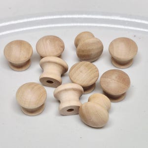 May include: A collection of unfinished wooden knobs with a rounded top and a cylindrical base. The knobs are light brown in color and appear to be made of natural wood. The knobs are likely for furniture or cabinet hardware.