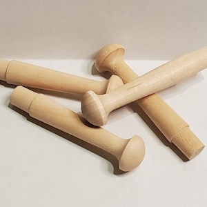 May include: Four unfinished wooden peg dolls, each with a rounded head and a cylindrical body. The pegs are light brown in color and appear to be made of natural wood. They are arranged on a white surface.