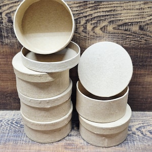 May include: A collection of round, beige paperboard boxes with lids. Some boxes are stacked, while others are open, revealing the interior. The boxes are arranged on a wooden surface, with a wooden background.