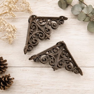 May include: Two dark brown cast iron shelf brackets featuring an intricate scrollwork pattern with curved lines and circular accents. Each triangular bracket has pre-drilled holes at the corners for mounting purposes and a textured metal finish.