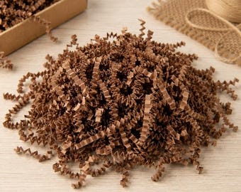 May include: A pile of brown crinkle-cut paper shred filler for gift packaging. The paper strips are thin, zig-zagged, and textured, forming a dense, loose mound. The material is a matte, earthy brown color, commonly used as protective cushioning.
