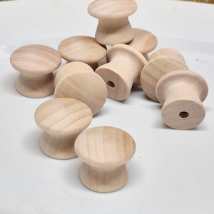 May include: A collection of unfinished wooden knobs with a mushroom-shaped top and a cylindrical base. The knobs are light brown and appear to be made of natural wood. They are scattered on a white surface.