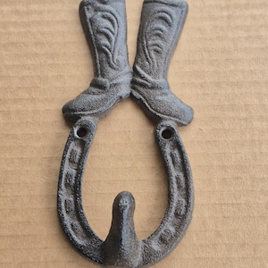 May include: A rustic, cast iron wall hook featuring a horseshoe and two cowboy boots. The boots are positioned above the horseshoe, creating a decorative and functional piece. The hook is finished in a dark, weathered color.