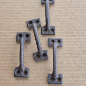 May include: Five antique-style, dark gray metal latches. Each latch has a rectangular base with two holes, a long, narrow stem, and a T-shaped top with two holes. The latches are arranged on a light brown surface.