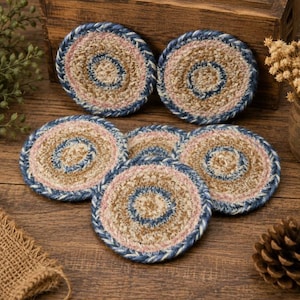 May include: A set of five circular coasters made from braided natural fiber rope. Each coaster features concentric rings of tan, light pink, and dark blue fibers, with a thick dark blue braided border around the outer edge of each circular piece.