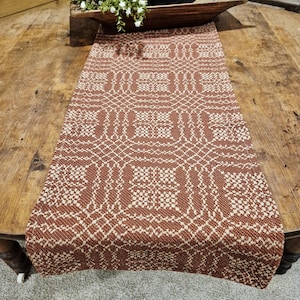 May include: A woven table runner with a geometric pattern in shades of brown and cream. The runner is displayed on a wooden table, showcasing its intricate design and texture. The runner adds a touch of rustic charm to any dining setting.