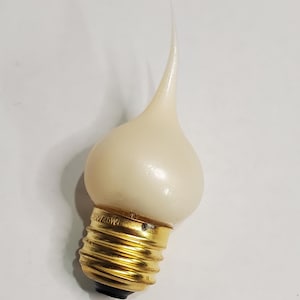 May include: A cream-colored light bulb shaped like a flame, with a pointed tip. The bulb has a gold-colored base with screw threads. The bulb is designed to look like a candle flame.