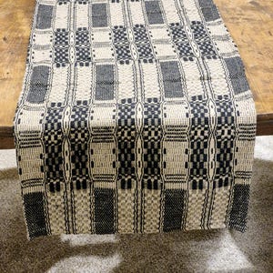 May include: Black and white woven table runner with a geometric pattern. The runner is made of a natural fiber and has a rustic look.