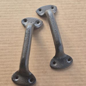 May include: Two cast iron door pulls with a dark gray finish. Each pull has a rectangular handle and two mounting plates with three holes each. The pulls are designed for easy installation and add a rustic touch.