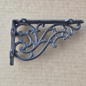 May include: A dark gray, cast iron shelf bracket with an ornate, scrolling design. The bracket has a right-angle shape with a flat top and a vertical support. The decorative elements include swirling patterns and small, circular details, perfect for a vintage or rustic decor.