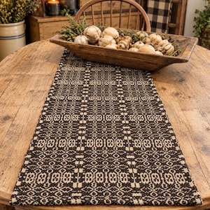 May include: A rectangular table runner featuring a dark brown and cream woven geometric pattern. Resting on the runner is an elongated wooden dough bowl filled with a collection of small, light-colored decorative gourds and green foliage.