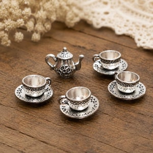 May include: A miniature silver-toned metal teapot and three matching teacups, each resting on a small saucer. The teapot features a decorative embossed pattern on its body, while the teacups and saucers have a simple, clean, metallic finish.