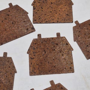 May include: Several small, rustic metal house cutouts with a weathered, brown finish. Each house has a rectangular shape with a pitched roof and a chimney. The cutouts are scattered across a white surface.