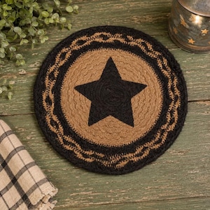 May include: A round braided jute mat featuring a natural tan center with a large black five-pointed star in the middle. The mat is bordered by a thick black braided ring and an outer edge of tan jute, creating a textured, circular design.