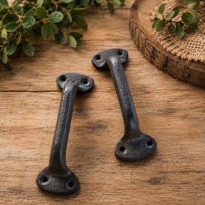 May include: Two heavy-duty, dark gray cast iron door handles with a textured, matte finish. Each handle features a curved grip and two circular mounting plates, each containing two pre-drilled screw holes for secure installation on doors or cabinets.