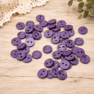 May include: A collection of small, round, purple buttons, each featuring two holes in the center. These circular fasteners are scattered across the surface, displaying a consistent solid color and a smooth, flat finish throughout the set.
