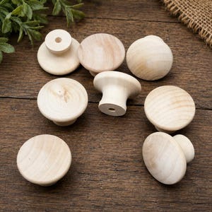 May include: A collection of seven unfinished light-colored wooden knobs with a smooth, rounded top surface and a tapered base. Each knob features a visible natural wood grain pattern and a central hole on the underside for mounting hardware.