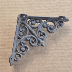May include: A decorative, cast iron shelf bracket with an ornate, scrollwork design. The bracket is dark gray and has a right-angle shape with pre-drilled holes for mounting. The design features swirling patterns and rounded accents, suitable for home decor.