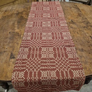 May include: A rectangular, woven table runner with a repeating geometric pattern in red and cream. The runner is displayed on a wooden table, showcasing its intricate design and texture. The runner's dimensions are not visible.