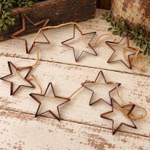 May include: A garland featuring six rusted metal star-shaped cutouts connected by a length of natural jute twine. Each star has a scalloped edge and a weathered brown finish, arranged in a loose, flowing line across the frame.