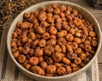 May include: A large, round, beige ceramic bowl filled with a dense collection of small, dried, orange-brown pumpkin-shaped fruits. The fruits have visible vertical ridges and stems, creating a textured, uniform appearance within the bowl.