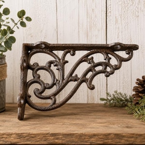 May include: A dark brown cast iron shelf bracket featuring an ornate, openwork scroll design with curved metal flourishes. The bracket has a flat top edge with two circular mounting holes and a vertical side edge with two additional mounting holes.