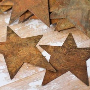 May include: Close-up of several rustic, star-shaped ornaments with a weathered, brown finish. The stars are arranged on a light-colored, textured surface, suggesting a vintage or farmhouse aesthetic. Some stars are stacked.