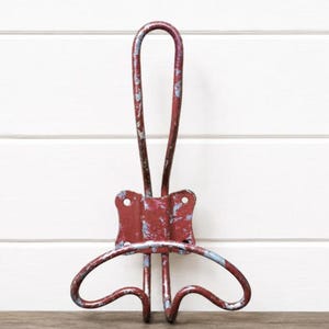 May include: A wall-mounted metal coat hook featuring a distressed red finish with visible gray underlayers. The hook has a tall, vertical loop at the top and two curved, wing-like arms at the bottom, with a central rectangular mounting plate.