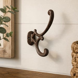 May include: A dark brown, textured cast iron wall hook featuring a double-pronged design with one long, curved upper arm and a shorter, curled lower arm. The mounting plate is oval-shaped with a visible screw hole for secure wall attachment.