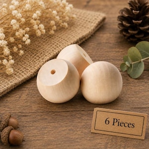 May include: A set of three light-colored, smooth, spherical wooden beads with a central hole, accompanied by a rectangular brown paper tag with the black printed text 6 Pieces. The beads have a natural, unfinished wood grain texture.