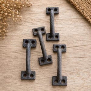 May include: A set of three dark gray, textured metal handles, each featuring a central bar and two rectangular mounting plates with two circular screw holes at each end, arranged in a vertical orientation on a light brown wooden surface.
