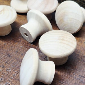May include: A collection of unfinished wooden knobs with a rounded top and a cylindrical base. The knobs are made of light-colored wood and are arranged on a dark wooden surface. The knobs are suitable for furniture or craft projects.