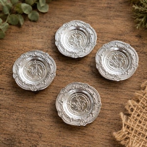 May include: Four small, round, silver-colored metal plates featuring a raised, intricate floral pattern in the center and a decorative, scalloped border with embossed circular details along the outer rim of each individual plate.