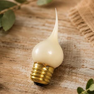May include: A flame-shaped light bulb with a matte, off-white glass finish and a metallic gold-toned screw base. The bulb features a distinct, elongated, and curved tip that tapers to a point, resembling the shape of a flickering candle flame.