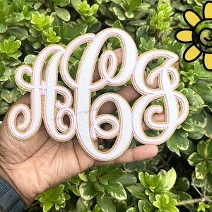 May include: A monogram in the letters A, B, and G, in a cursive font. The letters are white with a pink outline and gold glitter trim. The monogram is held in a hand against a background of green foliage.