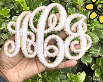 Può includere: Un monogramma decorativo con le lettere A, B e G in corsivo. Le lettere sono bianche con un contorno rosa e un bordo glitterato dorato. Il monogramma è tenuto in mano su uno sfondo di fogliame verde.
