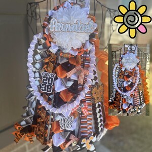 Puede incluir: Una gran flor de bienvenida hecha a mano en naranja, negro, blanco y plateado. La flor presenta cintas, lazos y una flor central con el nombre "Annabel". Incluye los textos "Class of 2038" y "HOCO 2025".