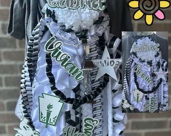 Può includere: Una mum di homecoming con nastri bianchi e verdi, con i nomi "Gabriel" e "Vivian". Include una stella, una moto e il testo "Hoco 2025".