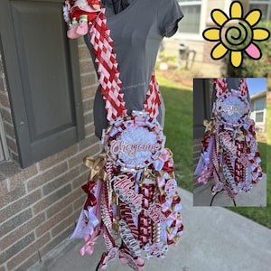 May include: A decorative mum with red, white, and gold ribbons, featuring the names "Cheyenne" and "Alberto". The mum includes a plush strawberry and is displayed on a mannequin. The design incorporates various textures and patterns.