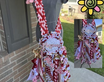 Può includere: Una decorazione tipo mum con nastri rossi, bianchi e dorati, con i nomi "Cheyenne" e "Alberto". La decorazione include una fragola di peluche ed è esposta su un manichino. Il design incorpora varie trame e motivi.
