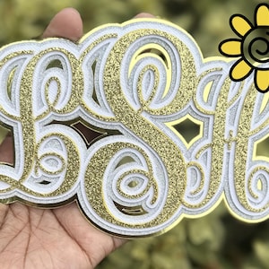 May include: A gold, white, and glitter monogram with the letters LSA. The monogram is held in a hand and has a decorative sunflower in the upper right corner. The letters are layered with a gold glitter center.