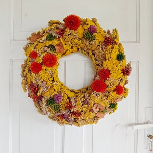 Puede incluir: Una corona de flores secas con flores amarillas, naranjas, rojas y moradas. La corona está colgada en una puerta blanca.