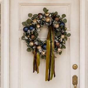 Pode incluir: Uma guirlanda circular com folhas de eucalipto verdes, detalhes em branco fosco e uma coleção de ornamentos esféricos em ouro metálico, bronze, azul-marinho e verde-escuro. Longas fitas fluidas em verde-oliva e marrom pendem da base.