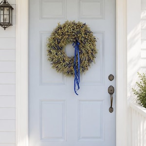 May include: A circular wreath composed of dried lavender sprigs, yellow billy button flowers, and green wheat stalks, accented with a long, flowing royal blue ribbon tied in a bow, hanging on a light blue six-panel front door with dark hardware.