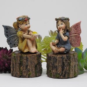 May include: Two fairy figurines with wings, sitting on tree stump bases. The fairy on the left is wearing a yellow dress and holding a yellow flower. The fairy on the right is wearing a blue dress and holding a green gem.