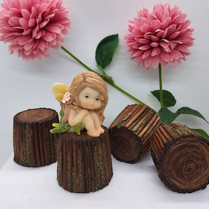 May include: A small fairy figurine with brown hair and a white flower on her dress sits on a brown log planter. The planter is surrounded by three other log planters, all with a brown bark texture. The background features pink flowers.
