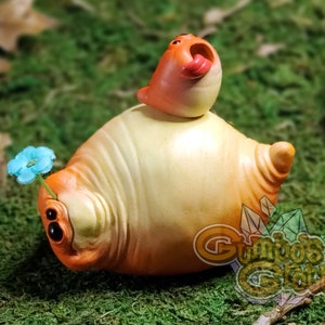 Può includere: Una scultura in ceramica dipinta a mano di una creatura di colore arancio chiaro e giallo con un piccolo uccello arancione appollaiato sulla schiena. La creatura ha un fiore blu sulla testa e una piccola coda arancione.