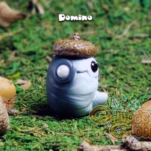 May include: A small, grey and white figurine with a brown acorn hat. The figurine has a single white eye and a small smile. The figurine is sitting on a bed of green moss.